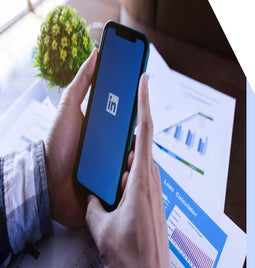 A person holds a smartphone displaying the LinkedIn logo, surrounded by papers with graphs and a small plant nearby.
