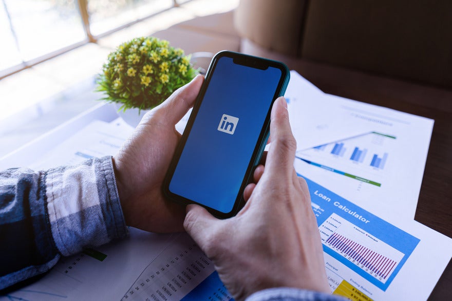 A person holds a smartphone displaying the LinkedIn logo, surrounded by papers with graphs and a small plant nearby.