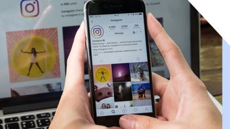 Hands hold a mobile phone showing Instagram's profile on Instagram, with a desktop behind also showing the same image