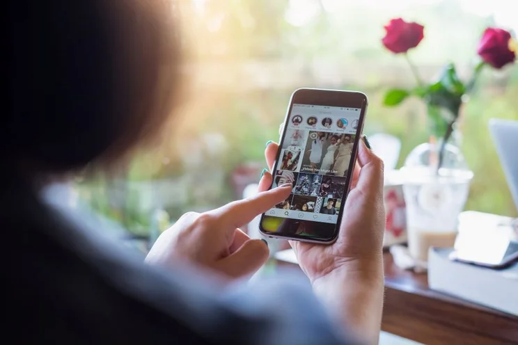 A person browses Instagram on their smartphone, with roses and a drink visible in the background, illuminated by natural light.