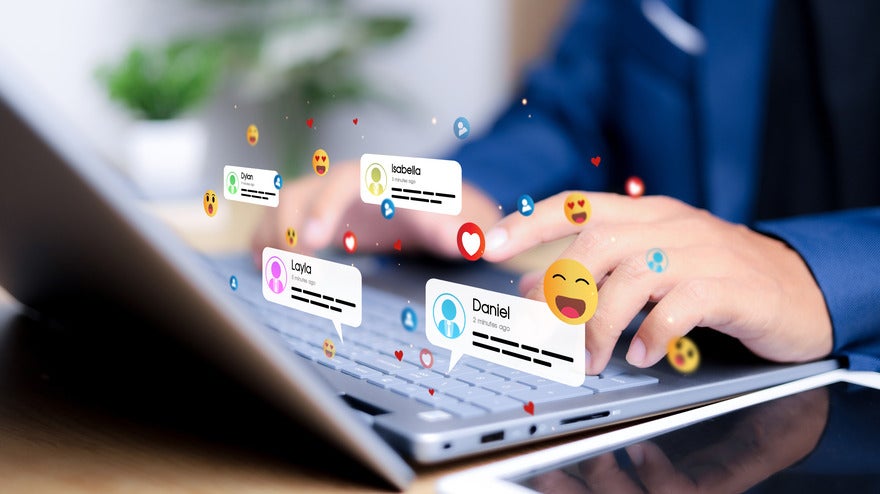 A person types on a laptop keyboard, surrounded by floating social media notifications and emojis representing interactions with friends.