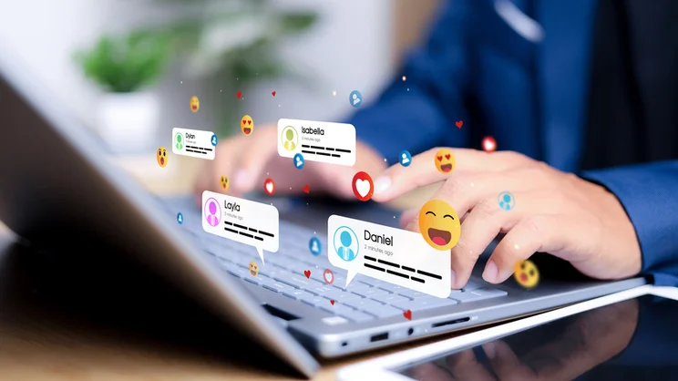 A person types on a laptop keyboard, surrounded by floating social media notifications and emojis representing interactions with friends.