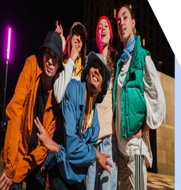 A group of five people in colorful streetwear poses playfully at night, with a vibrant pink light illuminating the background.