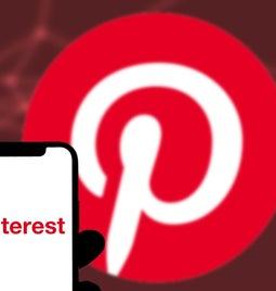 A hand holds a smartphone displaying the Pinterest logo, set against a blurred, red-backgrounded Pinterest symbol.