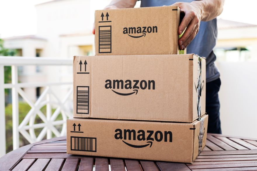 A person picks up stacked cardboard Amazon packages on a wooden table, with a blurred background of a sunny balcony.