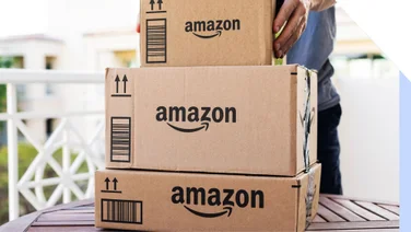 A person picks up stacked cardboard Amazon packages on a wooden table, with a blurred background of a sunny balcony.