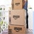 A person picks up stacked cardboard Amazon packages on a wooden table, with a blurred background of a sunny balcony.