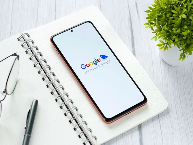 A smartphone displaying the Google Merchant Center logo rests on a notebook with glasses and a pen, beside a small potted plant.