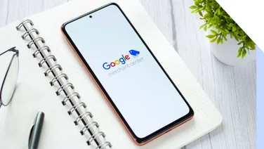 A smartphone displaying the Google Merchant Center logo rests on a notebook with glasses and a pen, beside a small potted plant.