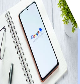 A smartphone displaying the Google Merchant Center logo rests on a notebook with glasses and a pen, beside a small potted plant.
