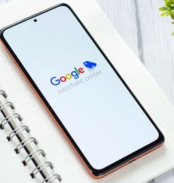 A smartphone displaying the Google Merchant Center logo rests on a notebook with glasses and a pen, beside a small potted plant.