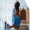 A person in a blue sweater sits at a desk with a laptop, tablet, and stationery, surrounded by clothing and shipping boxes.