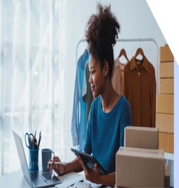 A person in a blue sweater sits at a desk with a laptop, tablet, and stationery, surrounded by clothing and shipping boxes.