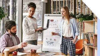 Three professionals collaborate in a modern workspace, discussing plans with a laptop, charts, and diagrams displayed on a board.