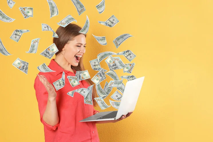 A person in a red shirt holds a laptop, surrounded by floating hundred-dollar bills against a bright yellow background.
