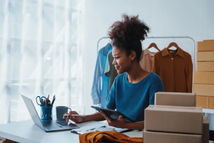 A person in a blue sweater sits at a desk with a laptop, tablet, and stationery, surrounded by clothing and shipping boxes.