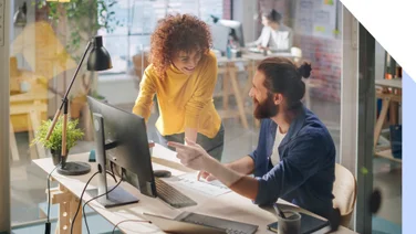 A collaborative workspace with two individuals discussing a project at a desk, surrounded by plants and modern office equipment.
