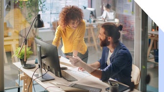 A collaborative workspace with two individuals discussing a project at a desk, surrounded by plants and modern office equipment.