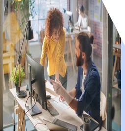 A collaborative workspace with two individuals discussing a project at a desk, surrounded by plants and modern office equipment.