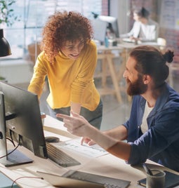 A collaborative workspace with two individuals discussing a project at a desk, surrounded by plants and modern office equipment.