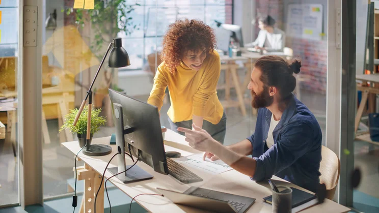A collaborative workspace with two individuals discussing a project at a desk, surrounded by plants and modern office equipment.