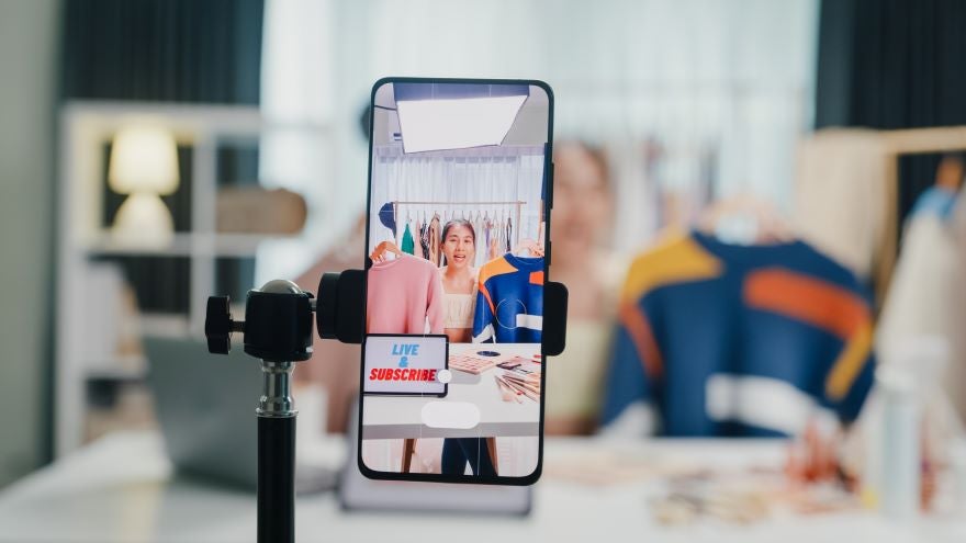 A smartphone on a tripod displays a clothing rack and makeup products in a bright studio, promoting a live streaming session.