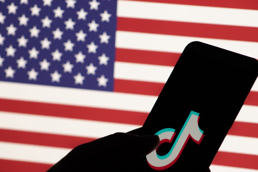 Silhouette of a hand holding a mobile phone with the TikTok logo on screen, the American flag is in the background