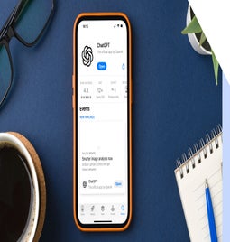 A smartphone displaying the ChatGPT app rating and features, surrounded by a coffee mug, glasses, and a notebook on a blue background.