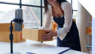 A woman in an apron lifts a cardboard box while preparing to record a video, surrounded by packing materials in a bright workspace.