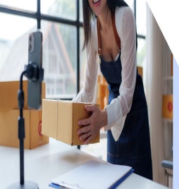 A woman in an apron lifts a cardboard box while preparing to record a video, surrounded by packing materials in a bright workspace.