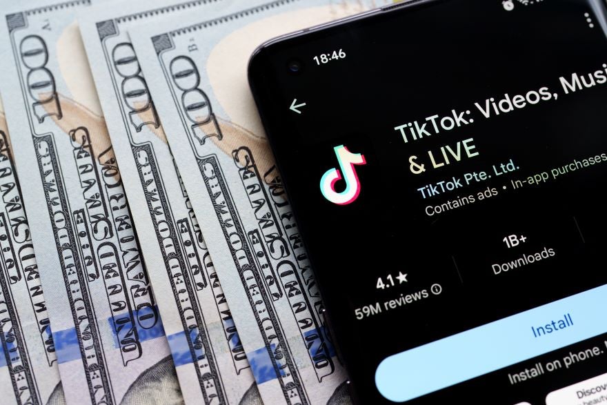 TikTok Money A smartphone displaying the TikTok app interface lies on a stack of hundred-dollar bills, highlighting digital media's connection to money.