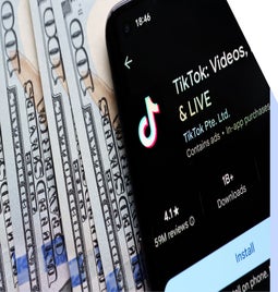 A smartphone displaying the TikTok app interface lies on a stack of hundred-dollar bills, highlighting digital media's connection to money.