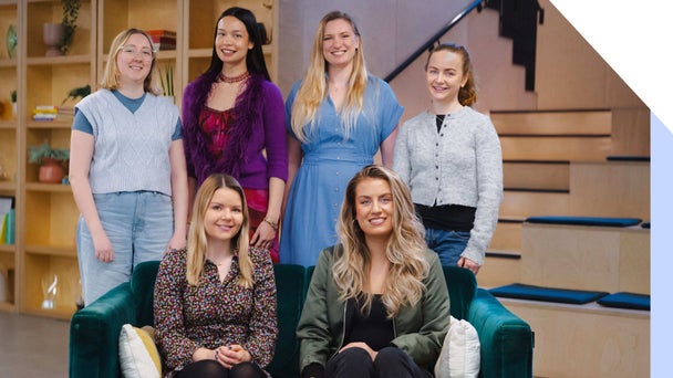 The Website Builder Expert team smiles at the camera, Heleana and Julia sat on a sofa and Emma, Holly, Lucy, and Annie stood behind them
