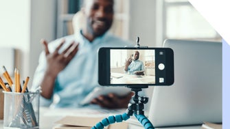 A person gestures while speaking to a smartphone on a tripod, surrounded by office supplies and paperwork in a bright workspace.