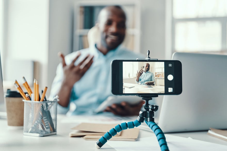 Social Media Trends November Intro A person gestures while speaking to a smartphone on a tripod, surrounded by office supplies and paperwork in a bright workspace.