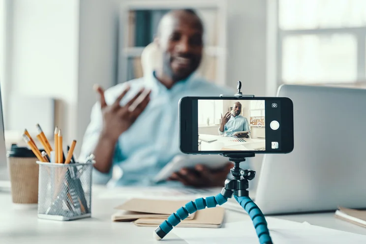 A person gestures while speaking to a smartphone on a tripod, surrounded by office supplies and paperwork in a bright workspace.