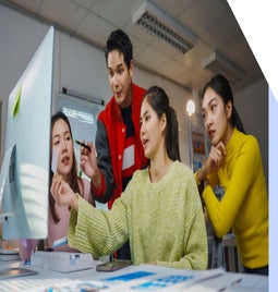 A group of four individuals collaborates around a computer, discussing ideas and examining documents in a modern office setting.