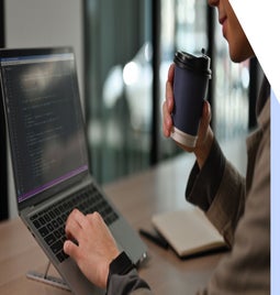 A person sat at a desk sipping coffee while editing code on a laptop