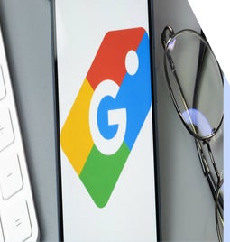 A smartphone displaying the Google logo, positioned beside a white keyboard and a pair of glasses on a gray surface.