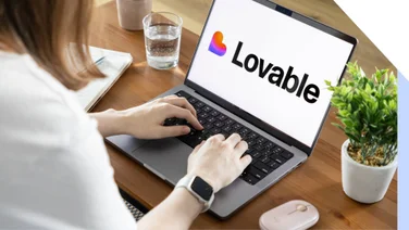 A person typing on a laptop displaying the "Lovable" logo, with a glass of water and a small plant on a wooden table.