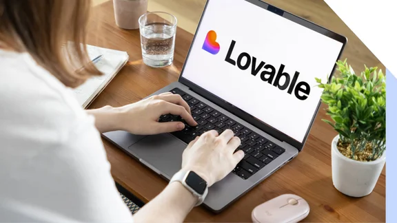 A person typing on a laptop displaying the "Lovable" logo, with a glass of water and a small plant on a wooden table.