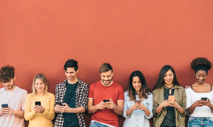 7 people leaning against a red wall looking down at their phones.