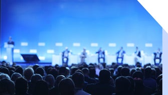 Large audience sat in chairs in front of a stage with speakers sat on chairs and a person stood to the side to moderate