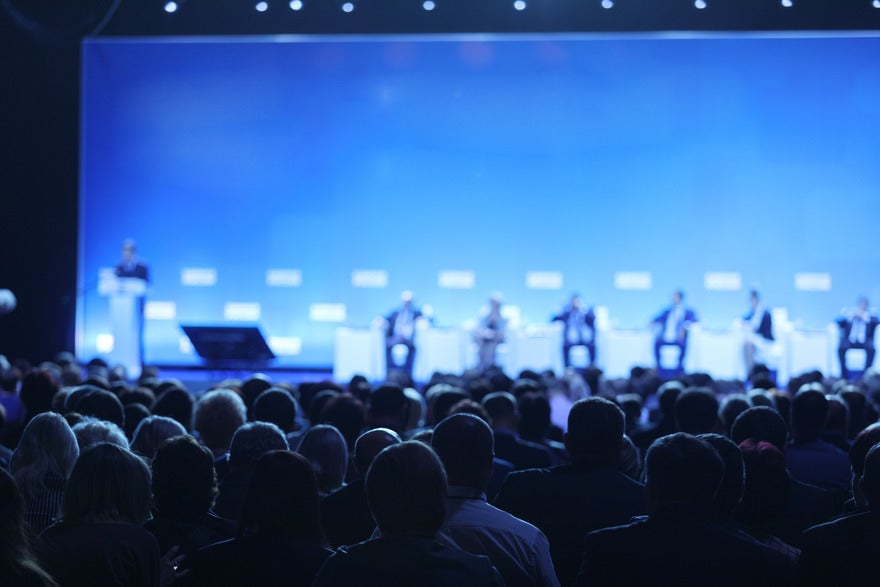 Large audience sat in chairs in front of a stage with speakers sat on chairs and a person stood to the side to moderate