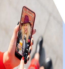 A hand holds a smartphone displaying a social media video, featuring a person with curly hair and a drink, against a brick background.