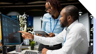 Two colleagues discuss a project at a modern office, focusing on a computer screen. One holds a tablet while pointing at the screen.