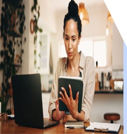 A person sits at a wooden table, holding a tablet, with a laptop and stationery in a cozy home office setting filled with plants.