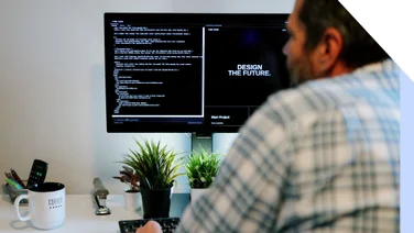 A person sits at a desk, programming on a computer with code on the screen, surrounded by plants and a coffee mug.