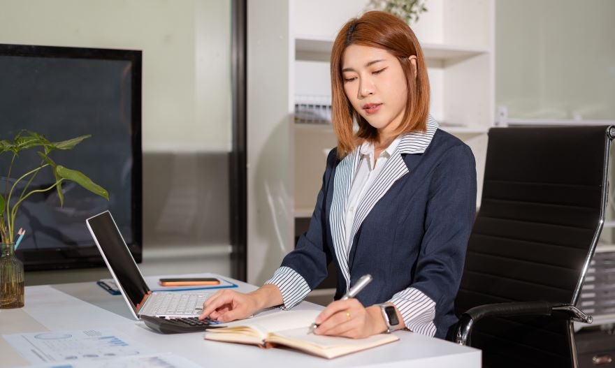 A person in a business outfit sits at a desk, using a laptop and writing in a notebook, surrounded by a modern office setting.