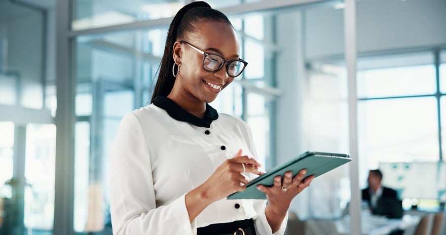 Businesswoman in a white blouse using a tablet in a modern office with glass walls and a blurred colleague in the background.
